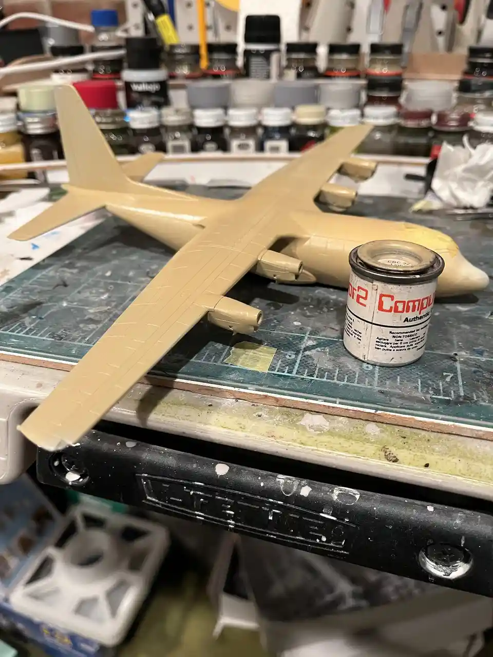 A beige model airplane sits on a cluttered hobbyist workbench next to a small paint can labeled 'Compul Authentic'. The…