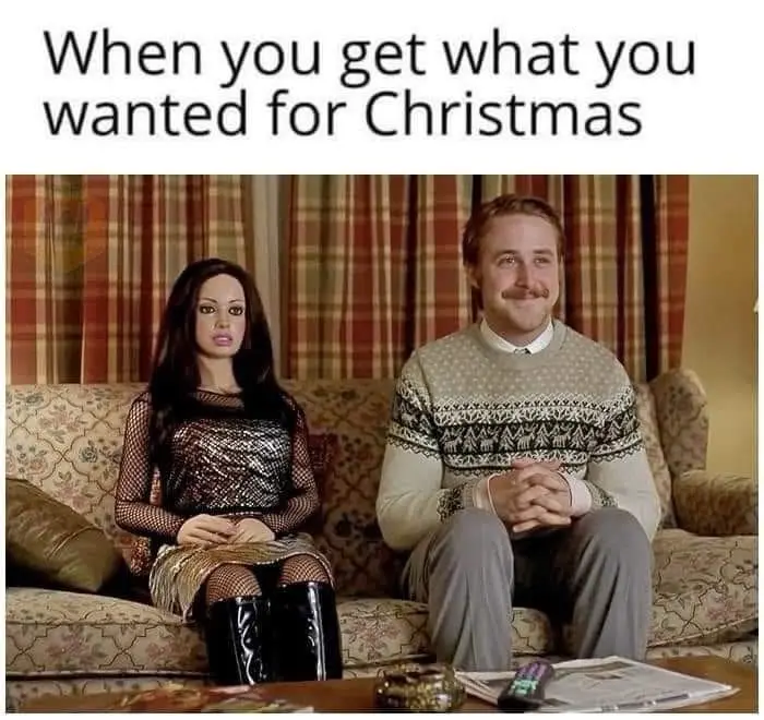 A man with a mustache wearing a patterned sweater sits on a floral couch next to a realistic-looking sex doll, both looking…