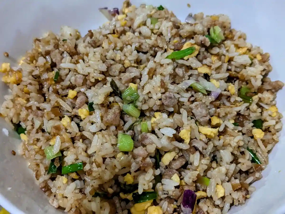 A close-up photo of a white bowl filled with fried rice containing visible grains, scrambled egg pieces, chopped green…