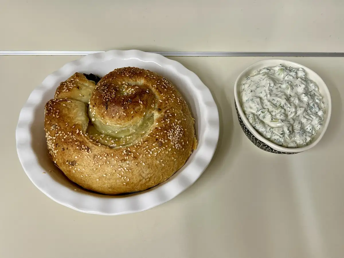 A golden-brown spiral pastry topped with sesame seeds sits on a white fluted plate, next to a small bowl of creamy white…