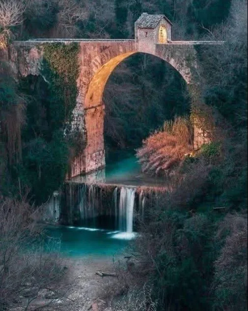 A scenic photo of an old stone arch bridge over a turquoise river with a small building on top, surrounded by trees. The…