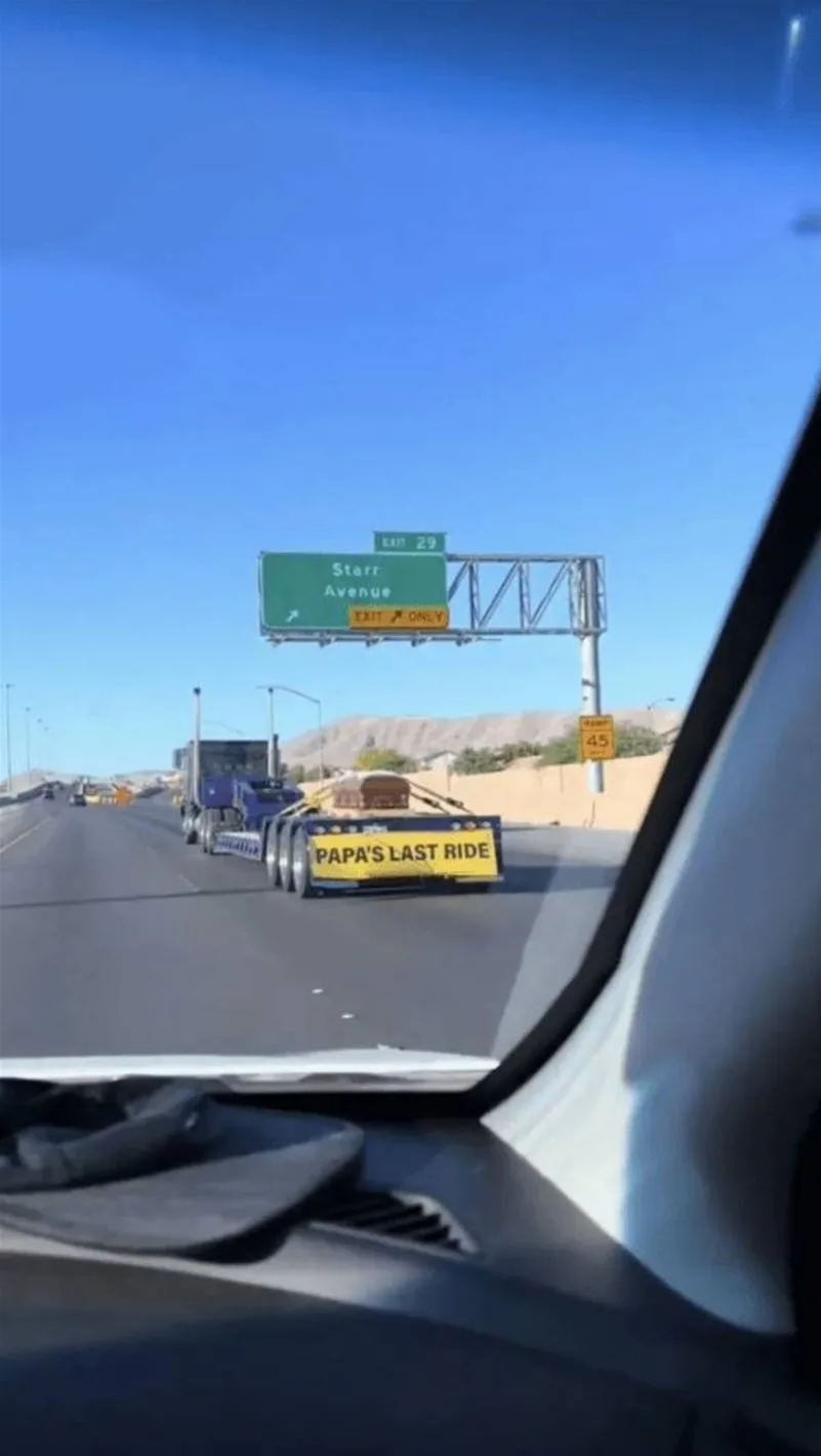 A photo taken from inside a car shows a blue semi-truck hauling a flatbed trailer with a yellow sign reading 'PAPA'S LAST…
