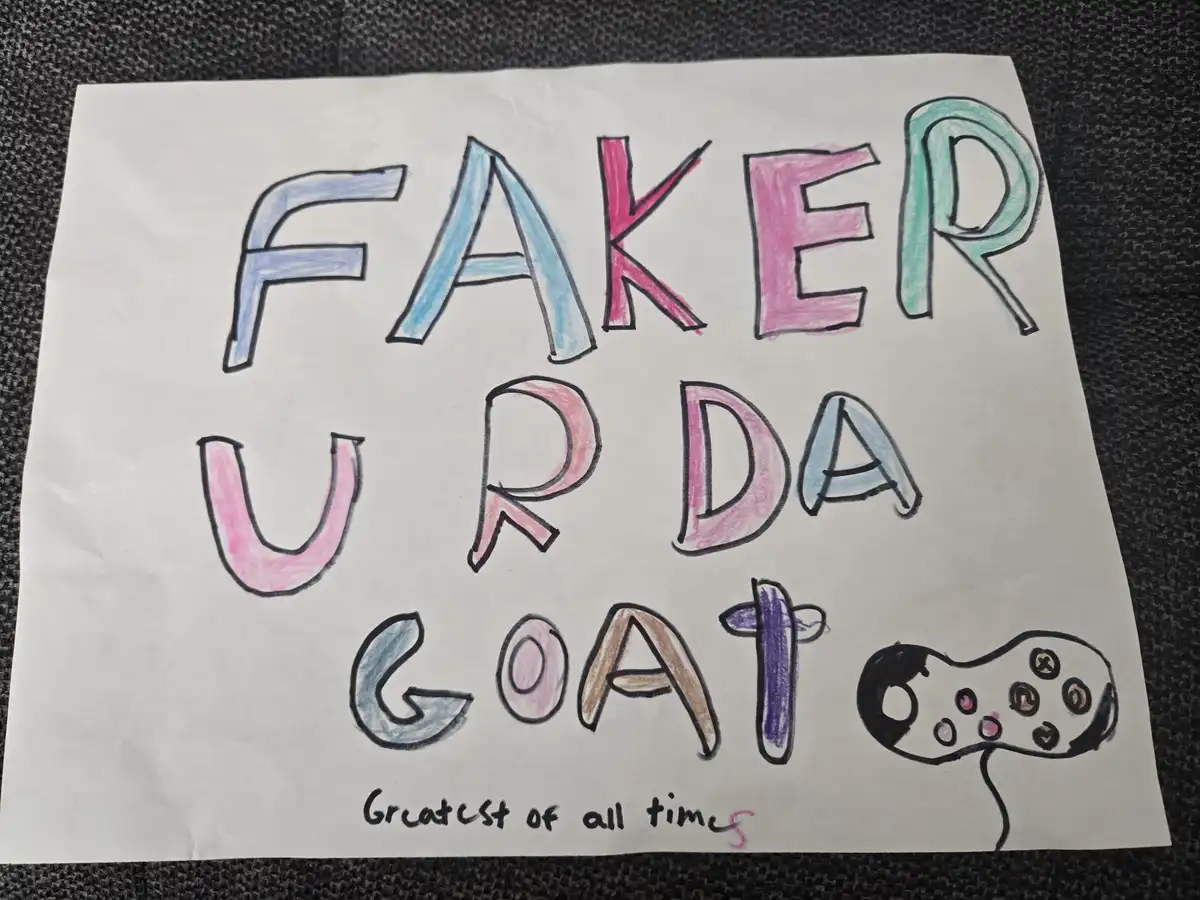 A hand-drawn sign on white paper with colorful crayon letters spelling 'FAKER U R DA GOAT' and a simple drawing of a game…