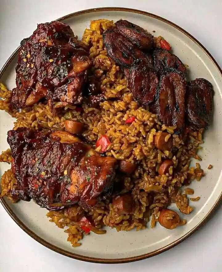 a top-down photo of a plate of jollof rice topped with glazed chicken pieces and fried plantains, with the text 'this is a…