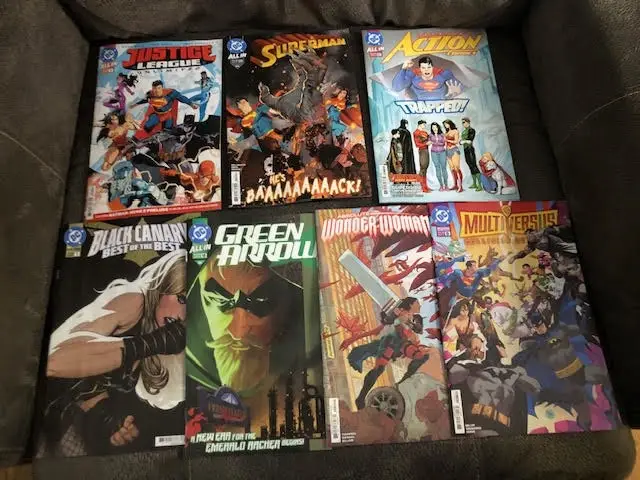 Six comic books are laid out on a dark couch, featuring Justice League, Superman, Action Comics, Black Canary, Green Arrow…