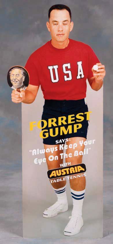 A man in a red USA t-shirt and black shorts holds a ping pong paddle with a photo of a smiling man on it and a ping pong…