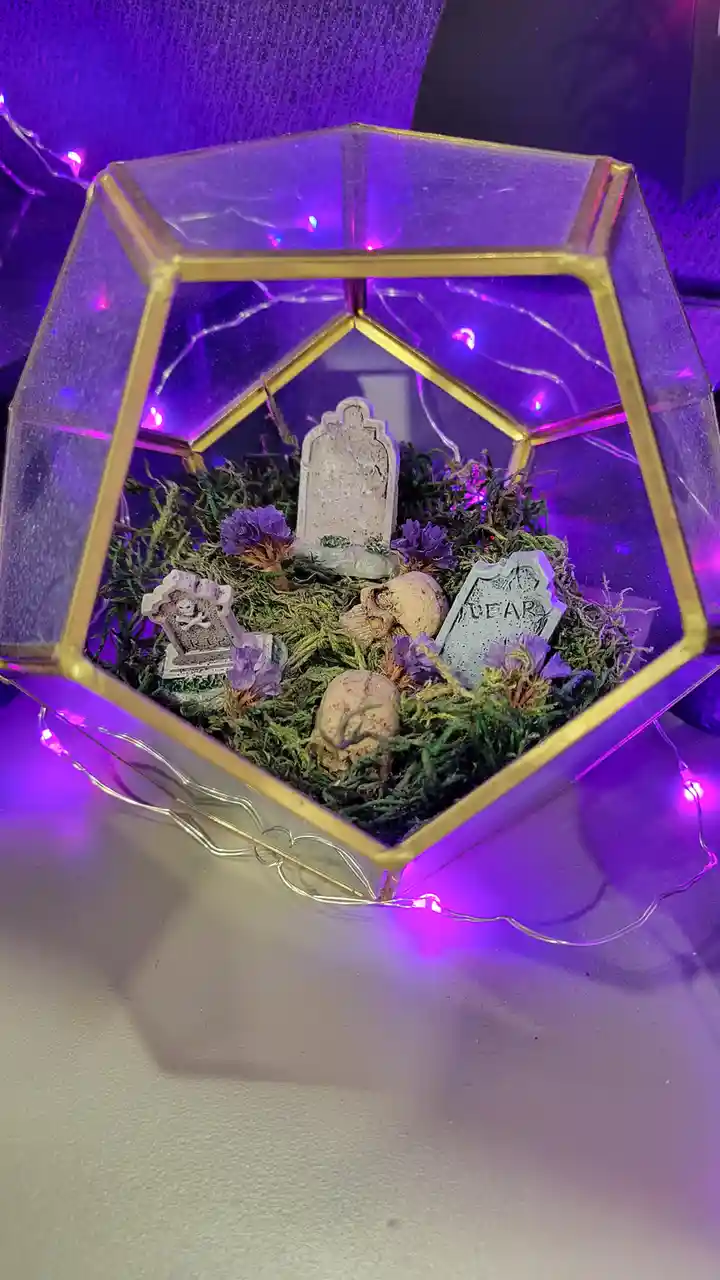 A hexagonal glass terrarium with gold trim contains a miniature graveyard scene with moss, purple flowers, skulls, and…