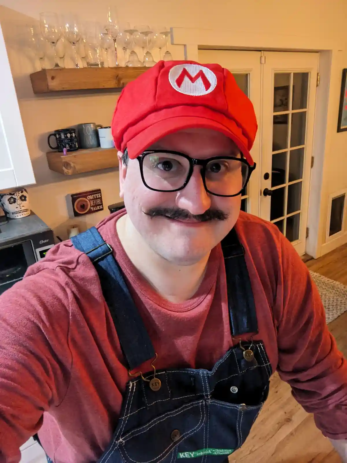 A man in a kitchen wearing a red Mario hat with a white 'M' logo, black-rimmed glasses, and a fake mustache, paired with…