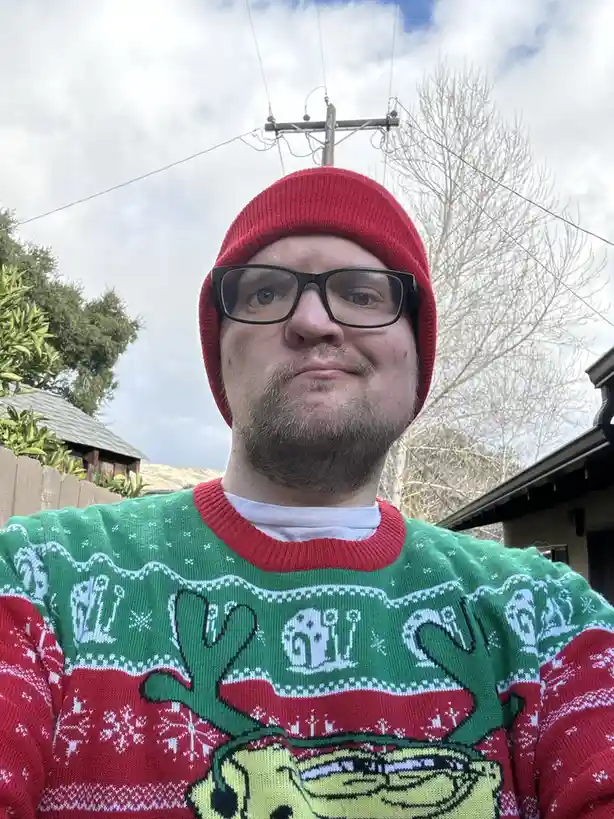 A man wearing a red beanie and black-rimmed glasses looks directly at the camera with a slightly annoyed or unimpressed…