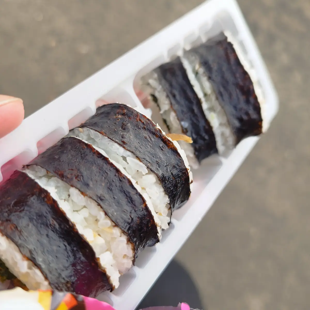 A close-up photo shows a hand holding a white plastic container with several pieces of sushi, each wrapped in dark nori…