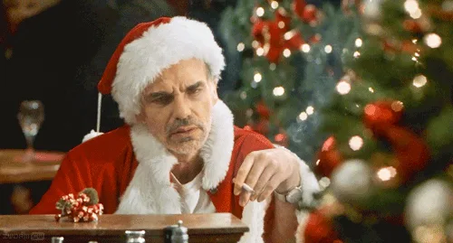A man dressed as santa claus sits at a table with a cigarette, looking deeply annoyed and unamused. Behind him, a decorated…