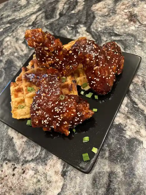 A black plate holds several glazed, sesame-seeded chicken wings resting atop golden-brown waffles, garnished with chopped…