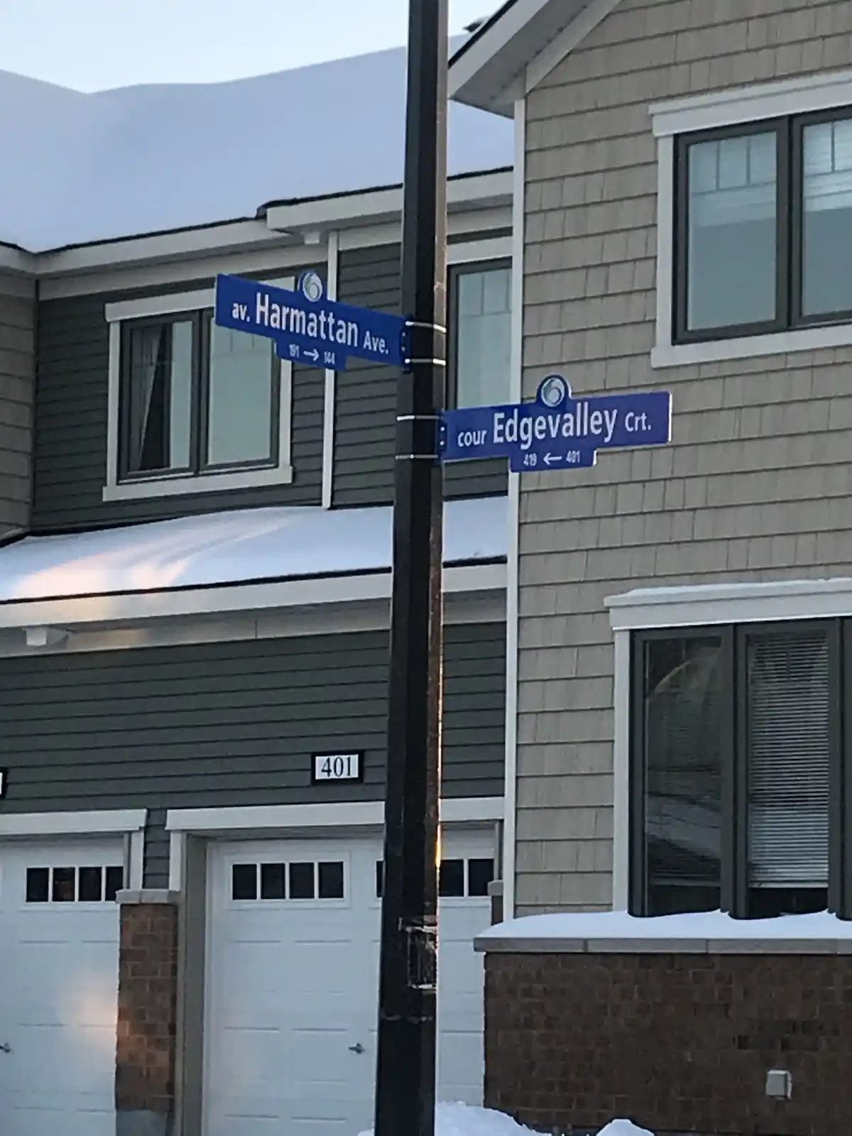 A snowy street corner shows two blue street signs on a pole: 'av. Harmattan Ave.' and 'cour Edgevalley Crt.', with house…