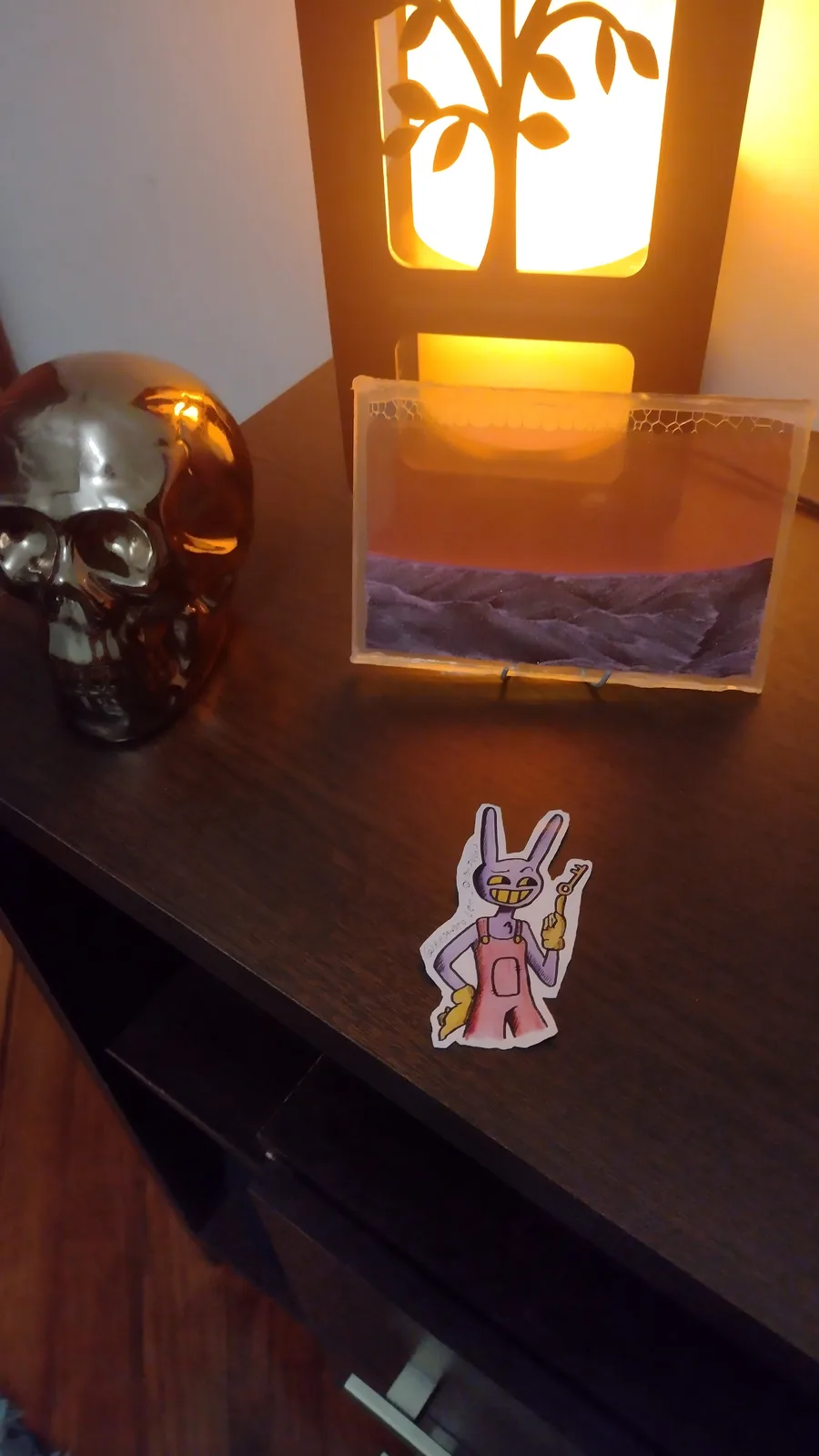 A photo of a dark wooden desk with a lit tree-shaped lamp, a metallic skull ornament, and a small framed desert landscape…