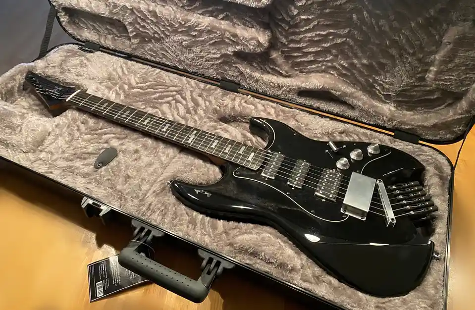 A black electric guitar with a unique, angular body and multiple pickups rests inside an open, plush-lined hard case. The…