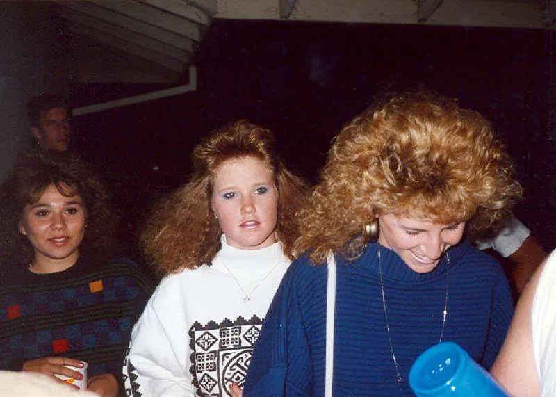 A grainy 1980s photo shows three young women at a party. The woman in the center, wearing a white turtleneck with a…