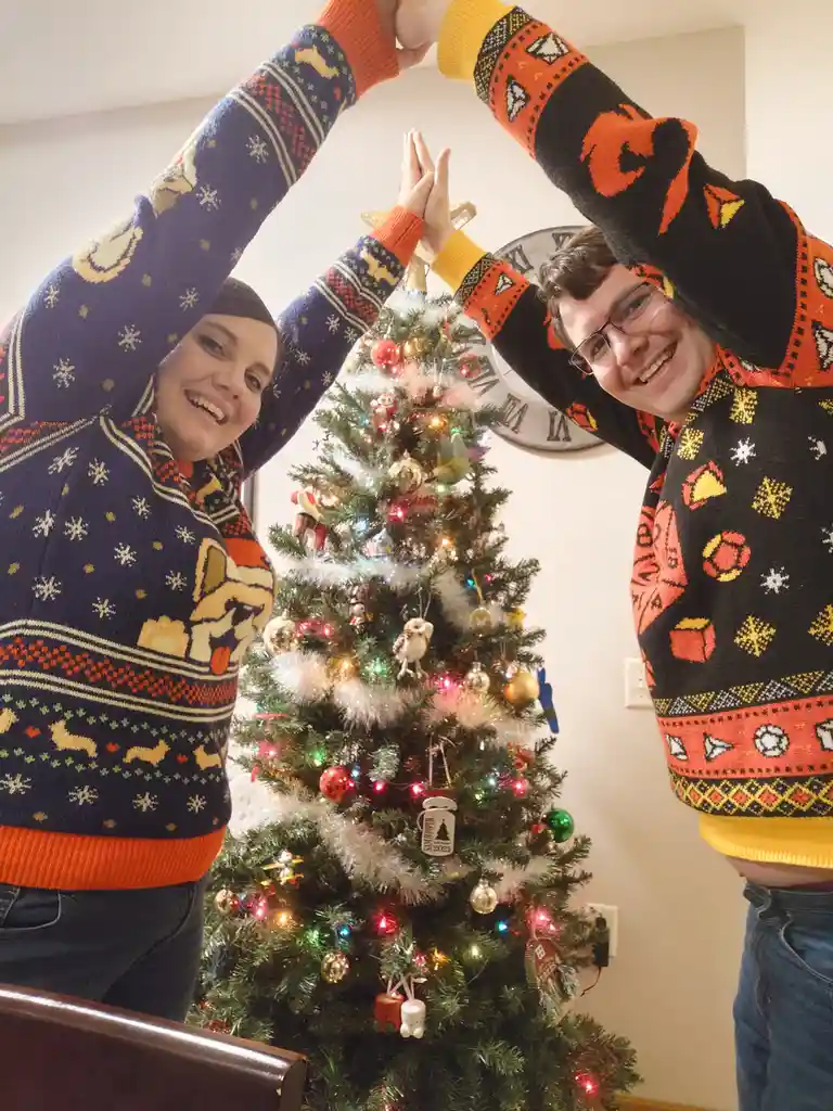 Two people in brightly patterned, festive Christmas sweaters stand on either side of a decorated tree, high-fiving at the…