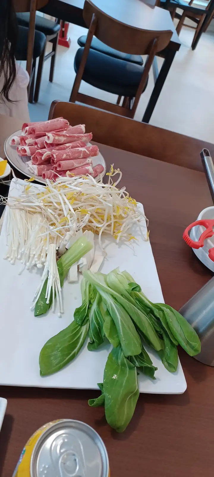 A photo of a hot pot meal setup with a plate of thinly sliced raw beef rolls, a white tray holding bean sprouts, enoki…