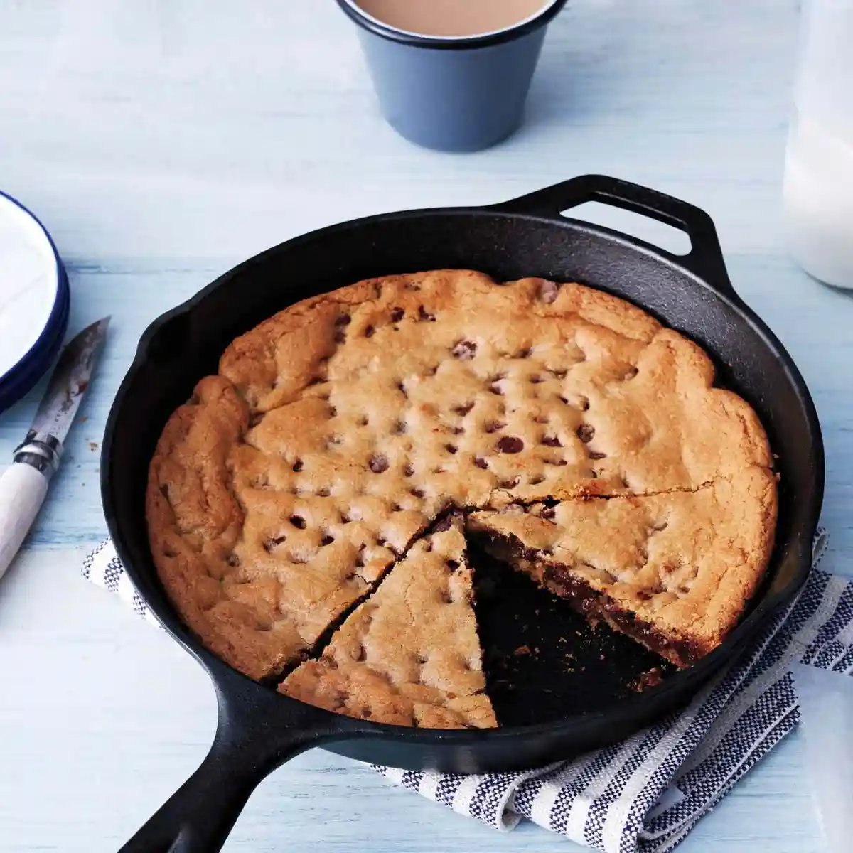 A large chocolate chip cookie baked in a black cast-iron skillet sits on a blue and white striped cloth, with a slice cut…