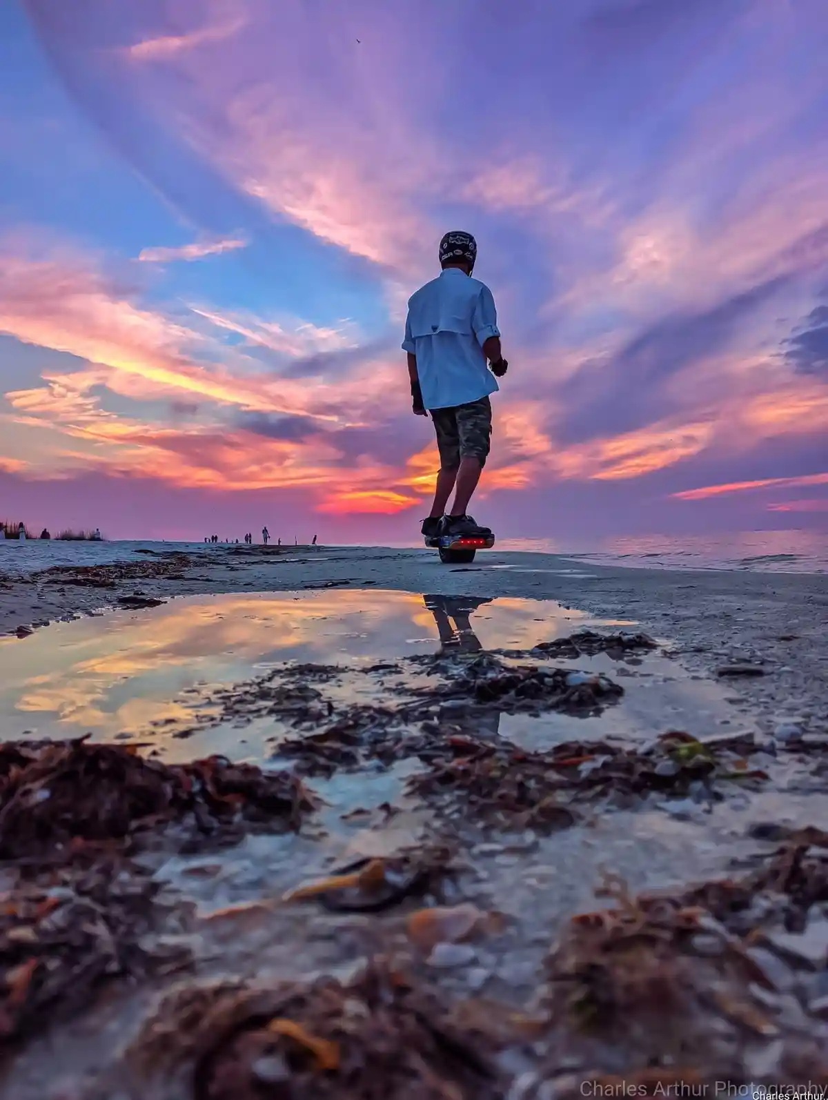 A person wearing a helmet and light blue shirt rides a hoverboard on a beach at sunset, with a vibrant pink and purple sky…