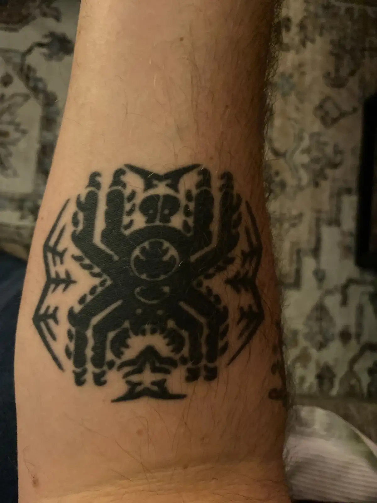 A close-up photo shows a black ink tattoo on a person's forearm, depicting a stylized spider with eight legs and a hexagonal…