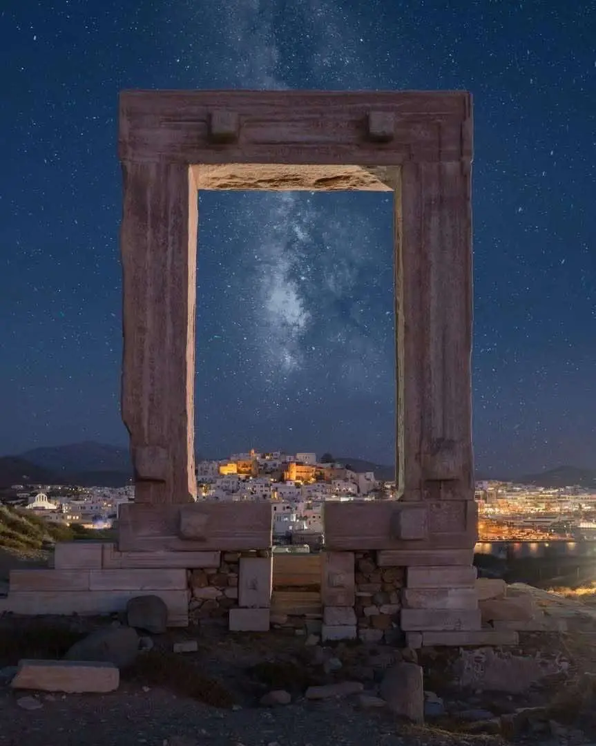 A large, ancient stone doorway frames a view of the Milky Way galaxy above a lit-up coastal town at night. The sky is filled…