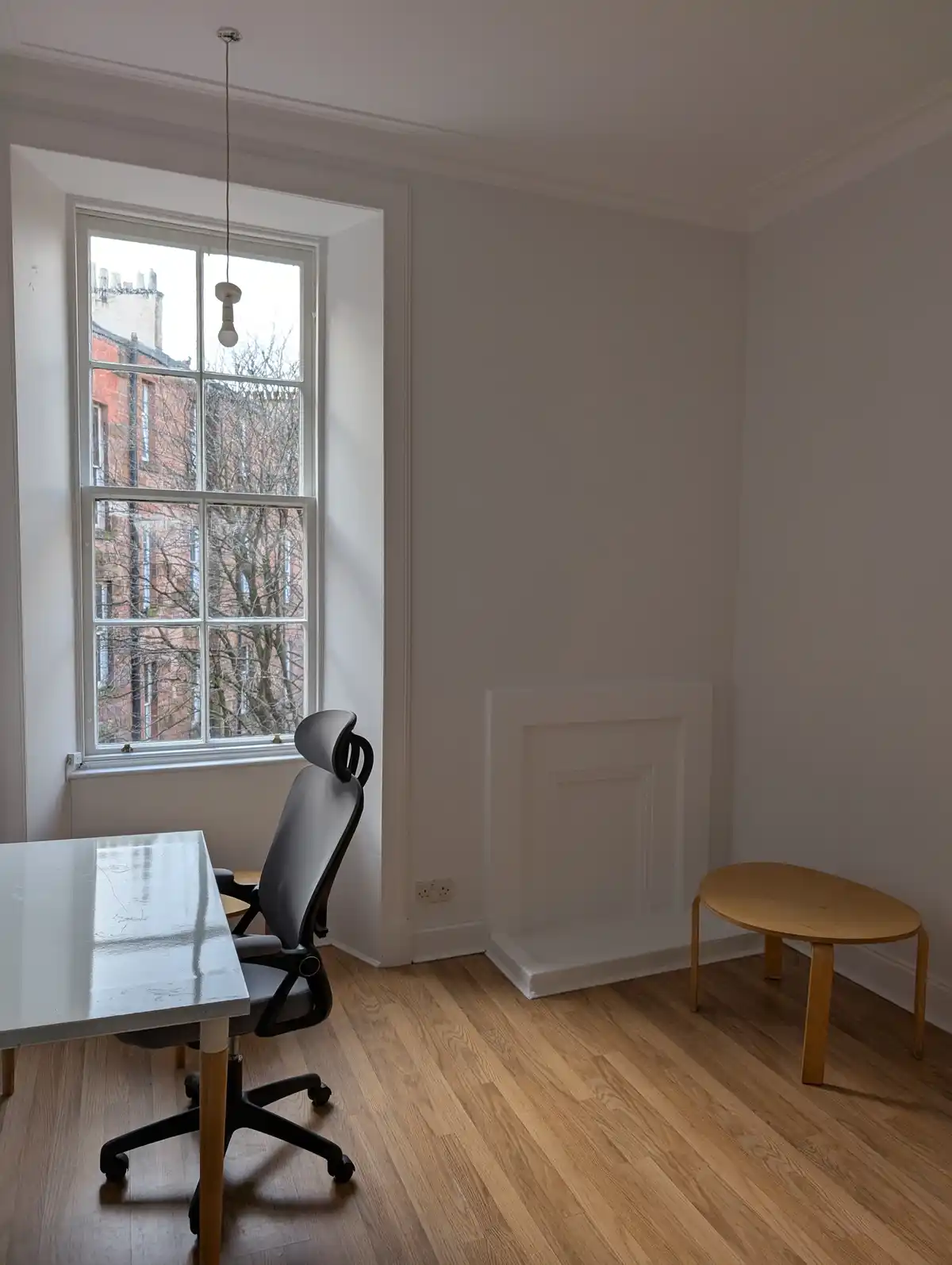 A minimalist, sparsely furnished room with light wood floors, white walls, and a large window showing brick buildings…