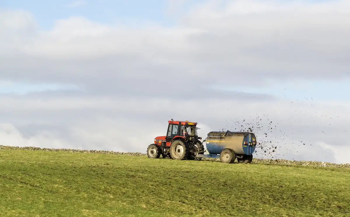 A red tractor pulls a blue tank across a grassy hill, kicking up dirt as it moves under a cloudy sky. The overlaid text…