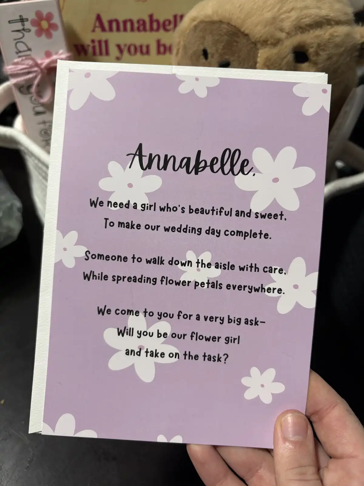 A hand holds a lavender-colored card with white flower patterns, addressed to 'Annabelle,' asking her to be a flower girl…