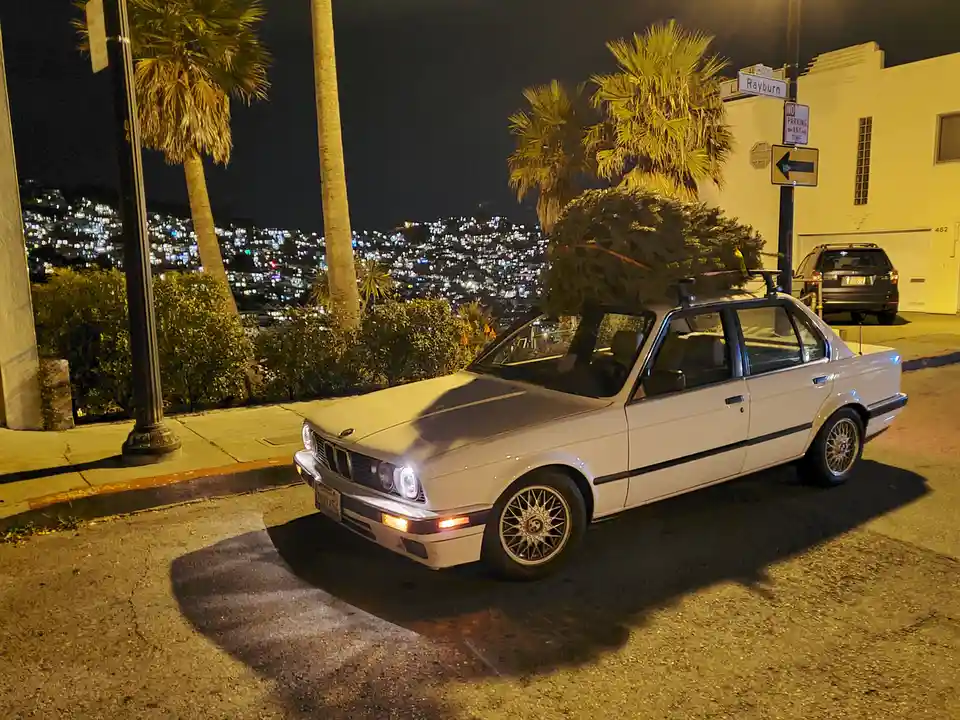 A white BMW E30 sedan is parked on a city street at night, with a large evergreen tree strapped to its roof rack. The…