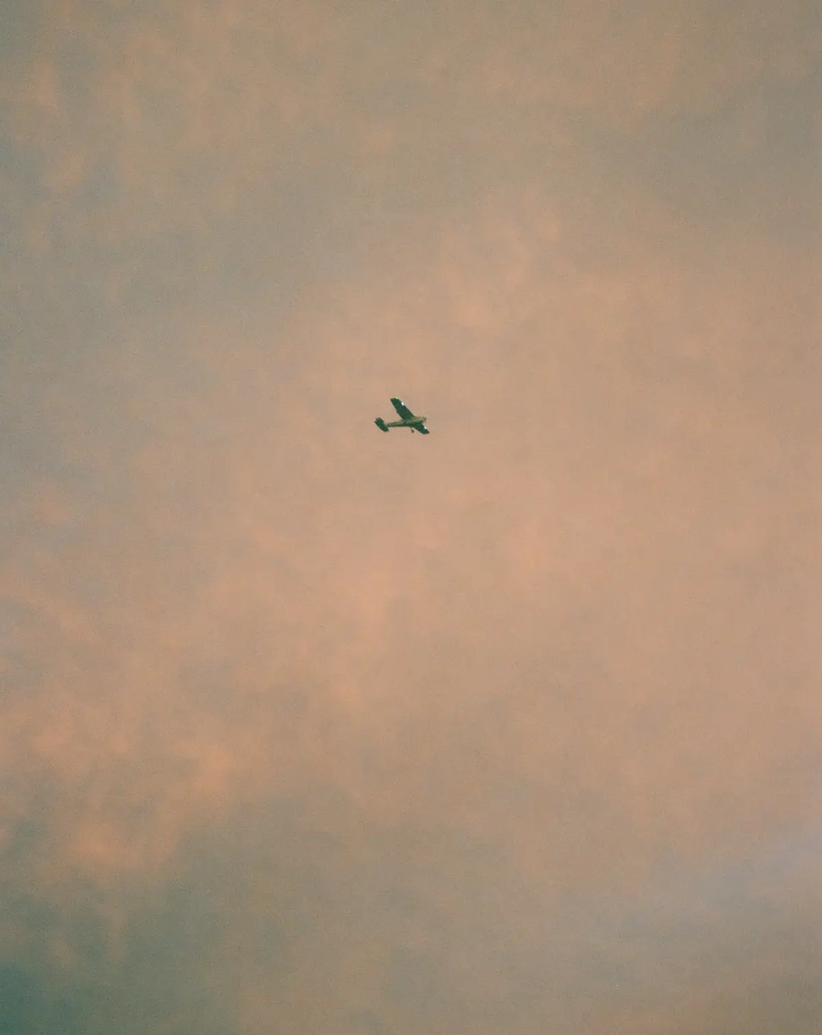 A small, dark-colored airplane is seen flying alone against a vast, hazy sky filled with soft pink and gray clouds. The…