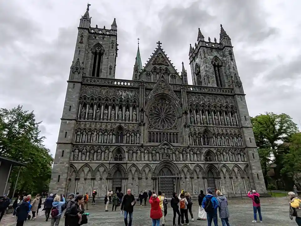 A large, ornate gothic cathedral with intricate stonework, statues, and spires under a cloudy sky, with tourists walking and…