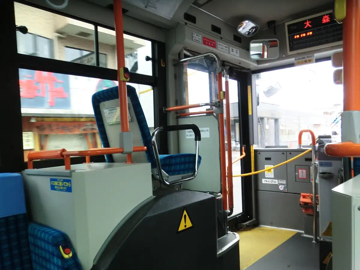 The image shows the interior of a Japanese bus, featuring blue plaid seats, orange handrails, and a digital display above…