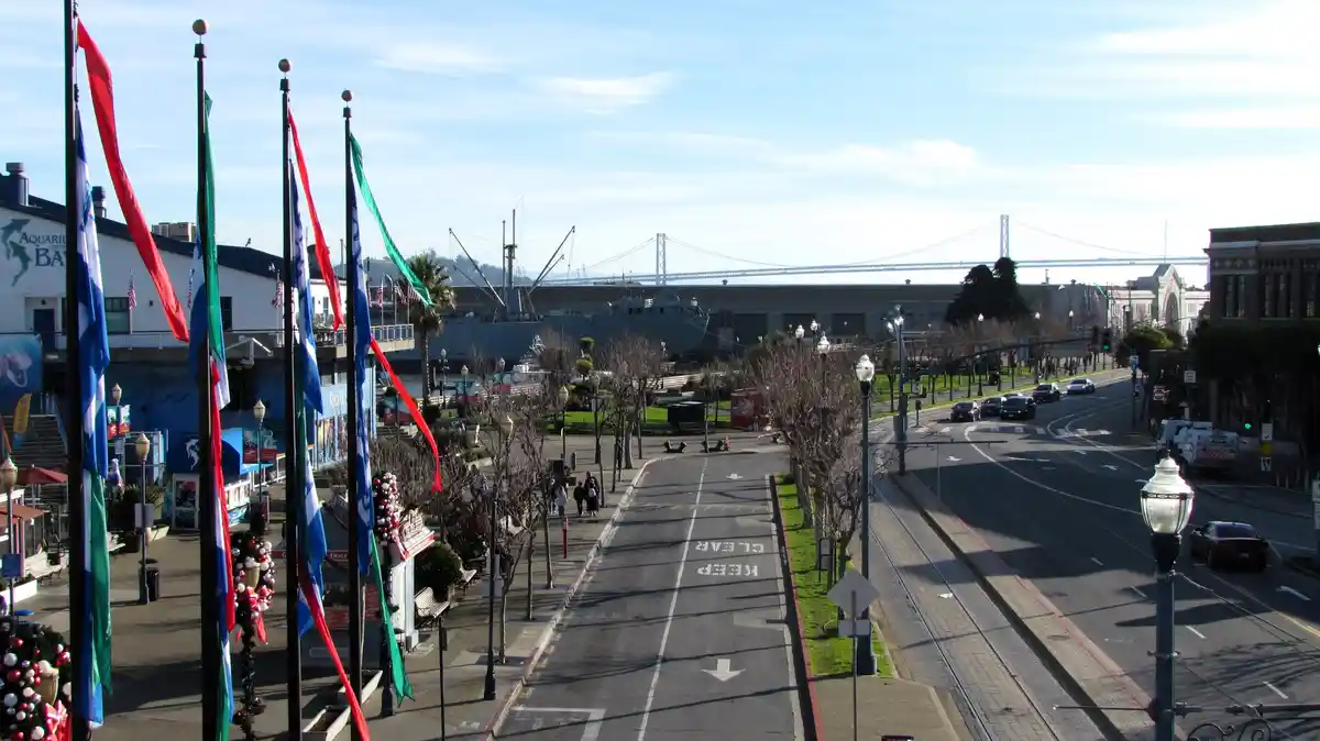 A wide-angle photo of a sunny waterfront street in San Francisco, featuring the Bay Bridge in the distance, the USS Golden…