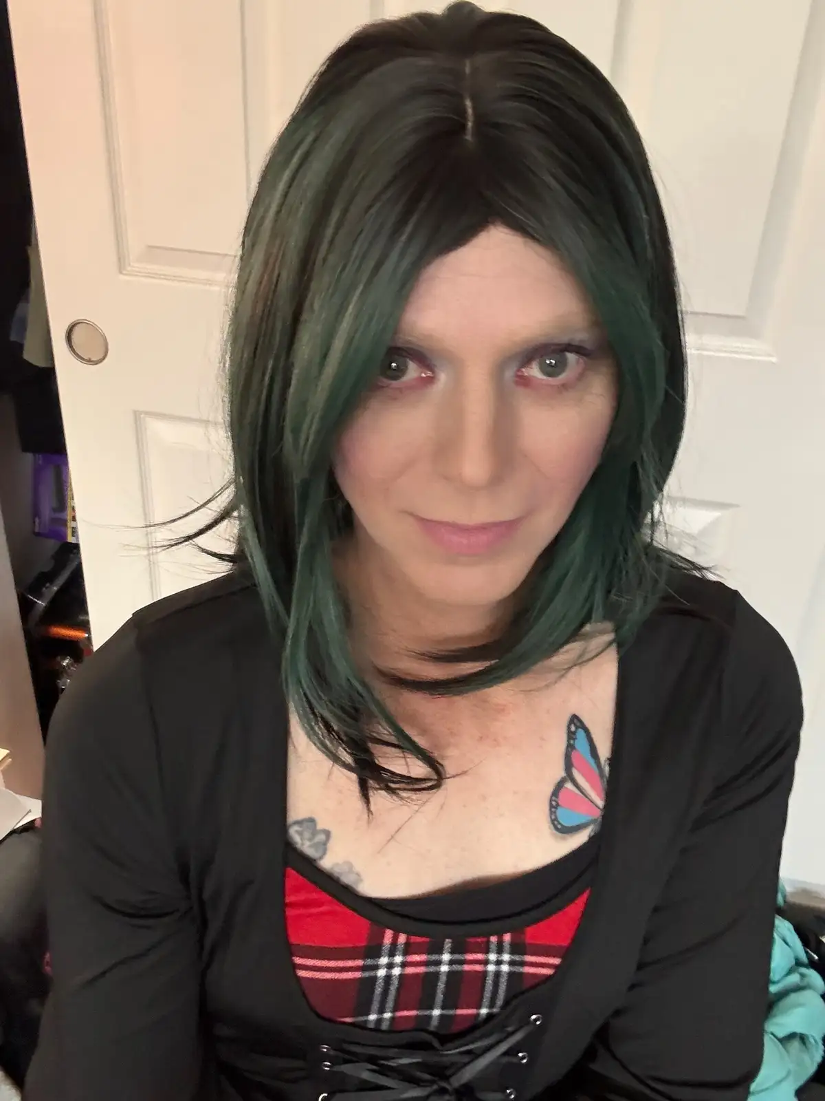 A person with dark green hair and heavy eye makeup stares directly at the camera with a subtle smirk. They wear a black…