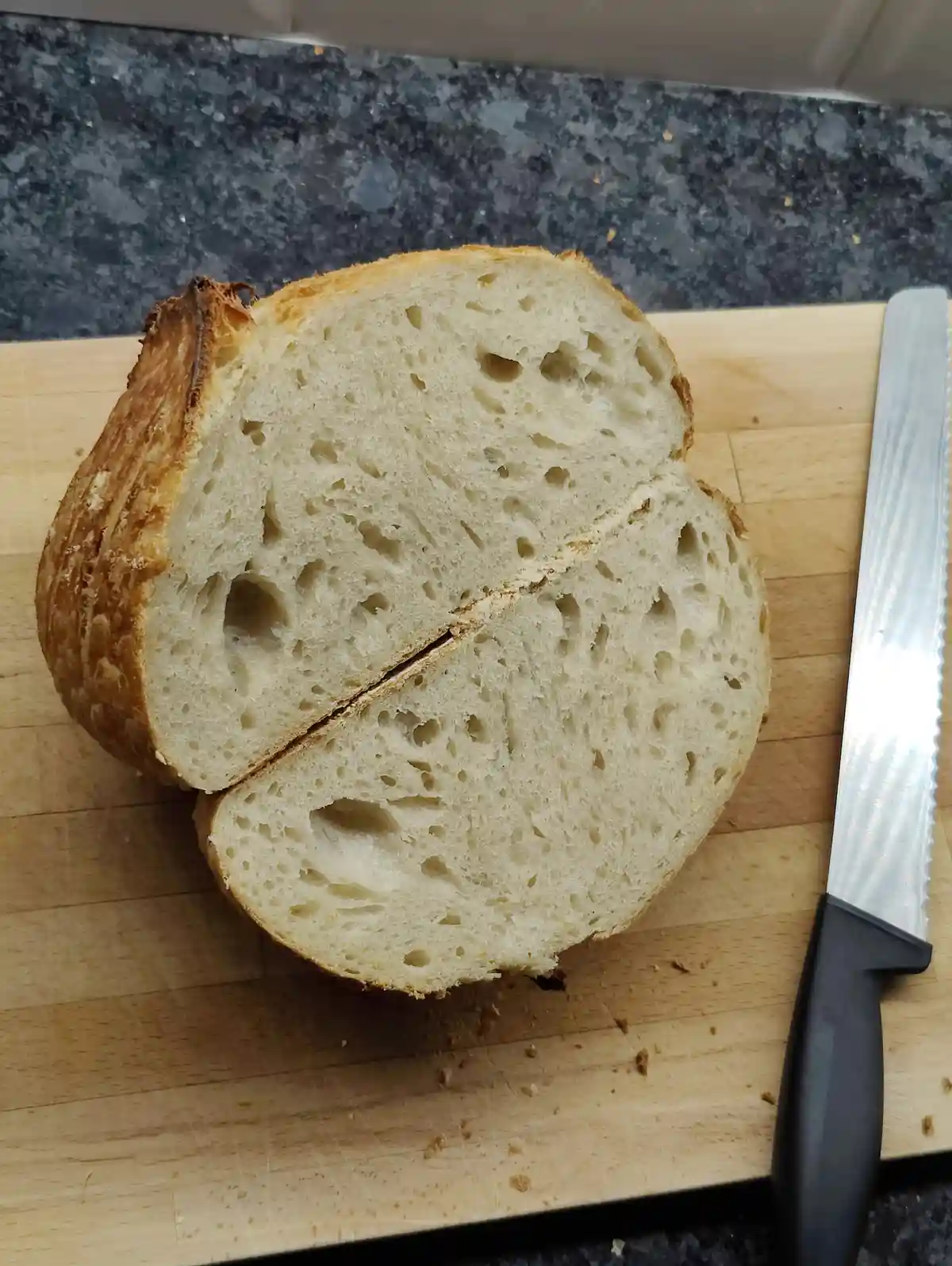 A loaf of artisan bread, sliced in half and resting on a wooden cutting board, has a crack down the middle that resembles a…
