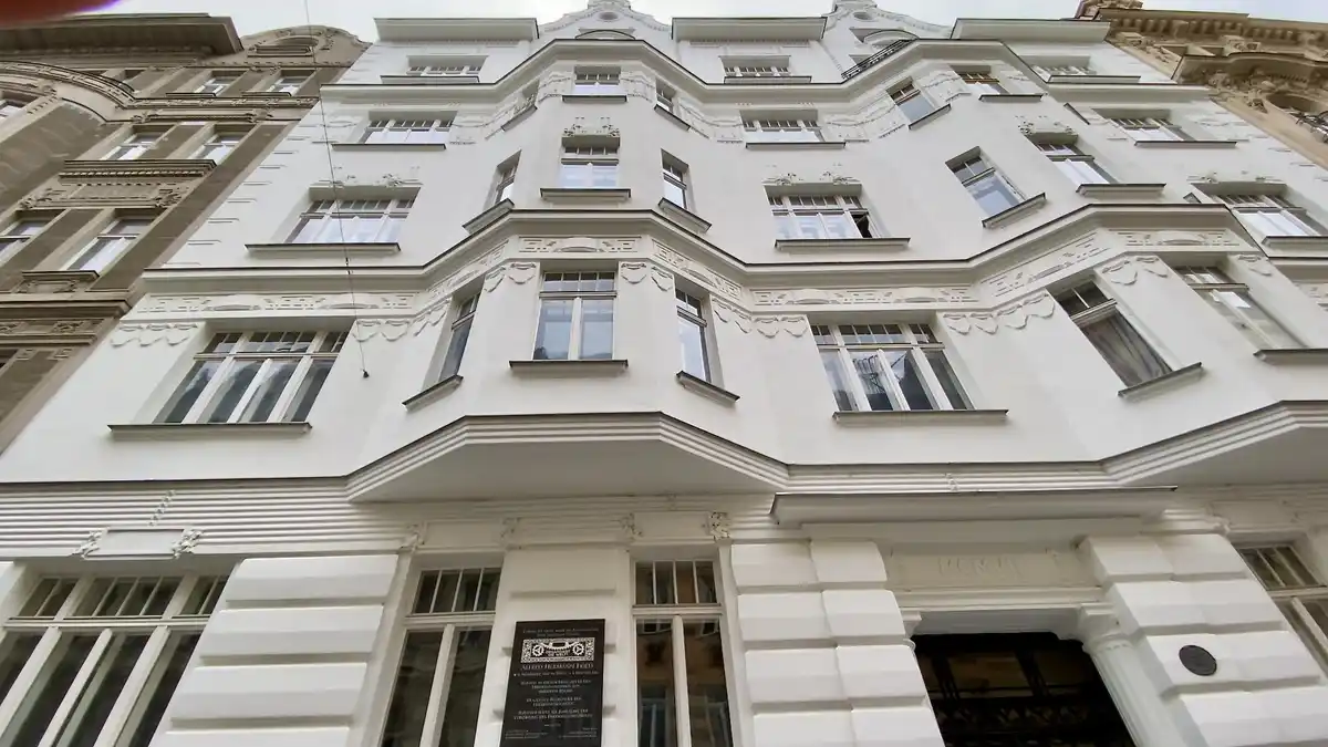 A low-angle photo shows the ornate, white facade of a multi-story European building with many windows and decorative…