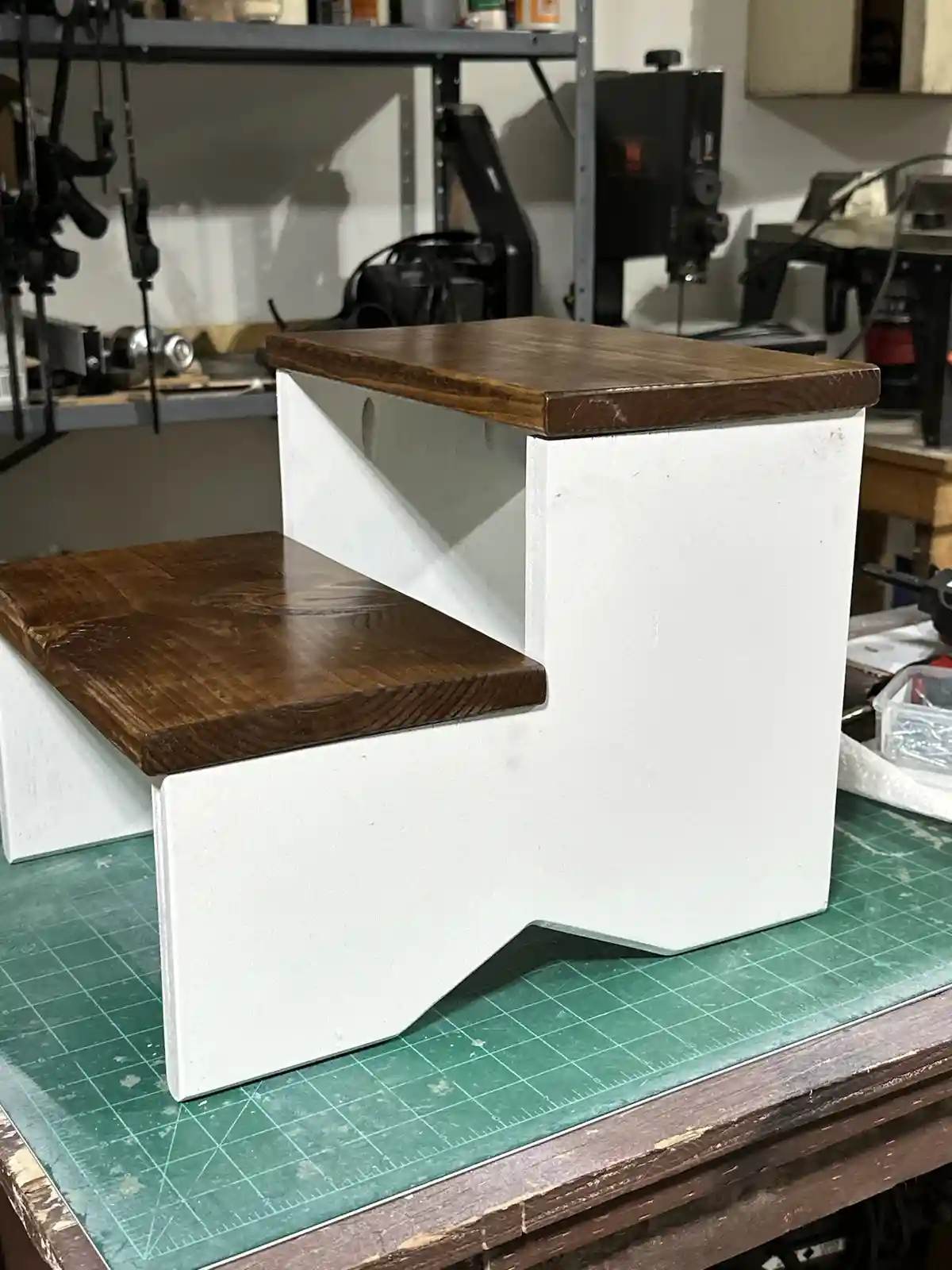 A handmade two-step stool with white painted sides and dark wood tops sits on a green cutting mat in a workshop. The…