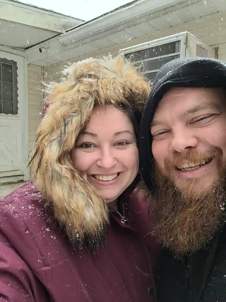 A selfie of a smiling man and woman outdoors during a snowfall, both wearing winter jackets and hoodies with snowflakes on…