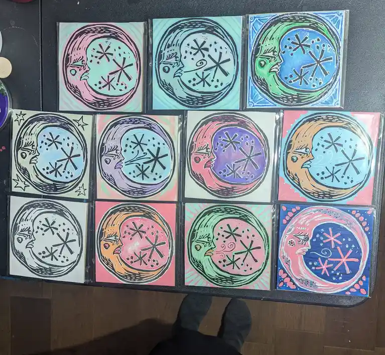 An overhead photo shows twelve small, colorful, hand-painted ceramic tiles arranged in a 3x4 grid on a black surface. Each…
