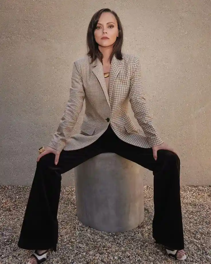A woman with shoulder-length brown hair sits on a cylindrical concrete block outdoors, wearing a beige and black checkered…