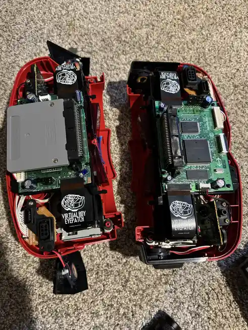 Two disassembled red Nintendo Virtual Boy consoles lie side by side on a carpet, exposing their internal circuit boards…