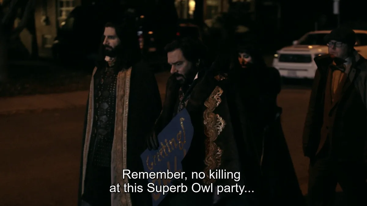A nighttime street scene shows four men in costumes walking; one holds a blue sign that reads 'Greetings!' while white text…