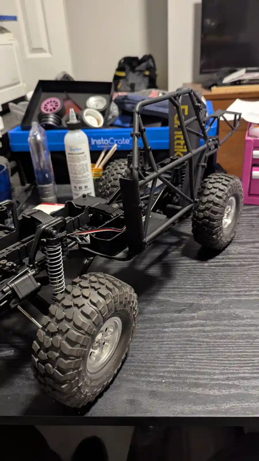 A partially assembled black remote-controlled off-road vehicle with large knobby tires sits on a dark wooden table…