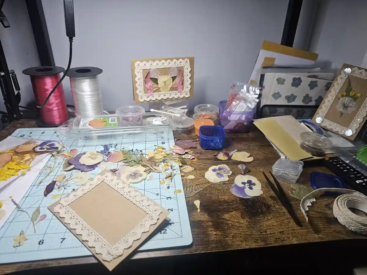 A cluttered craft desk is filled with dried flowers, lace-edged cards, spools of ribbon, tweezers, and various crafting…