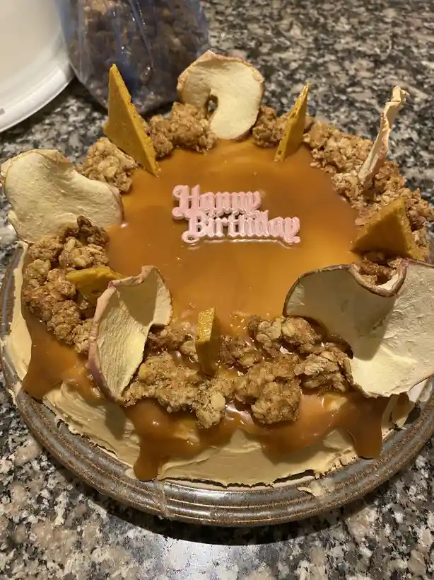 A close-up photo of a birthday cake decorated with caramel sauce, crumble topping, and dried apple slices arranged around…