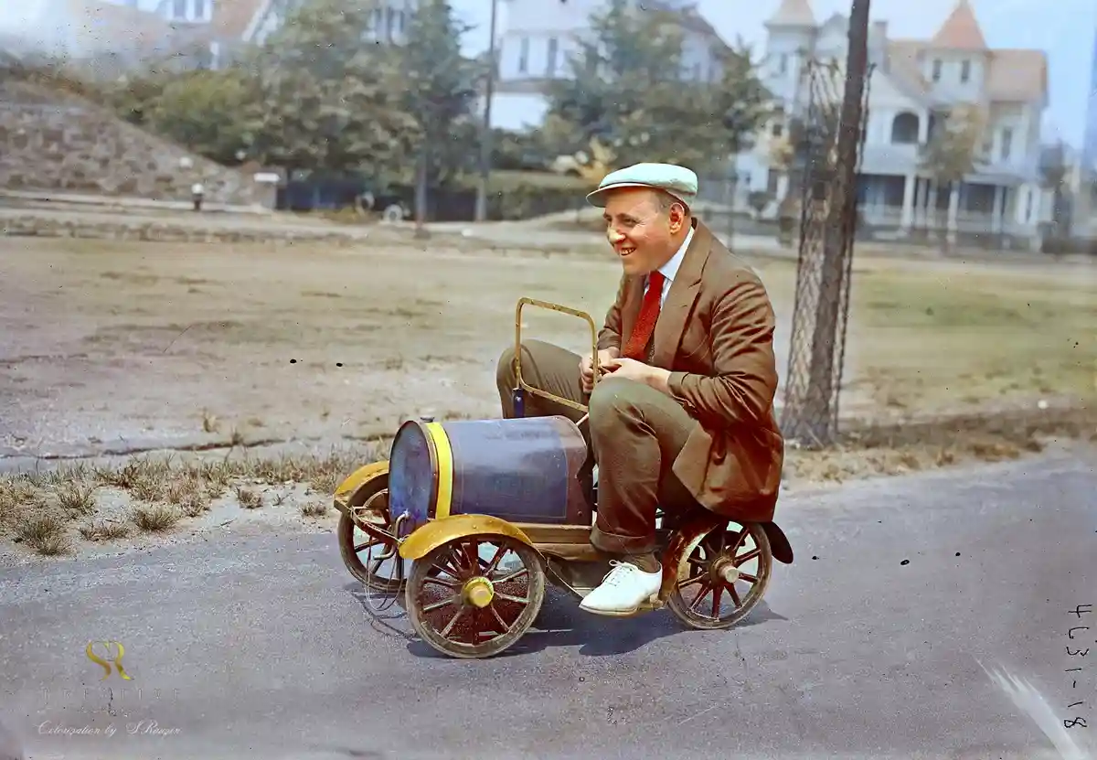 A man in a suit and cap sits smiling on a very small, rudimentary, barrel-shaped vehicle with spoked wheels, looking off to…