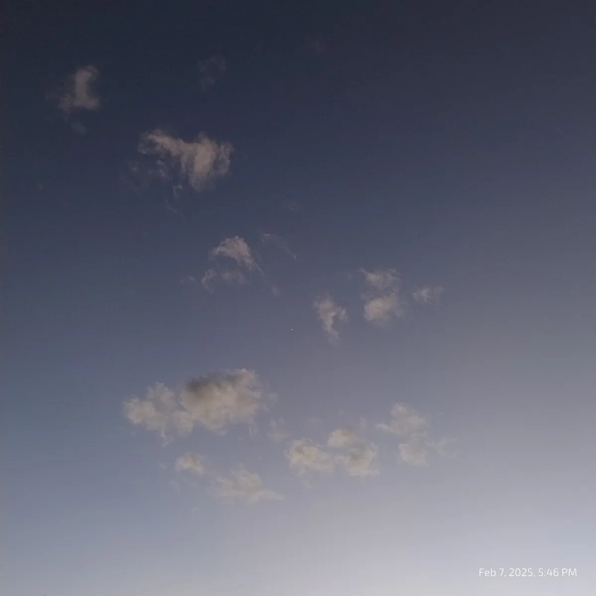 A photo of a twilight sky with scattered white clouds against a gradient blue background, featuring a small bright dot…