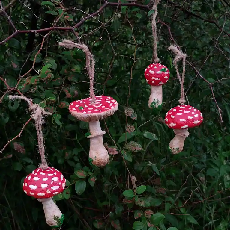 Four hand-painted red-and-white spotted mushroom ornaments hang from twine tied to thorny branches against a backdrop of…