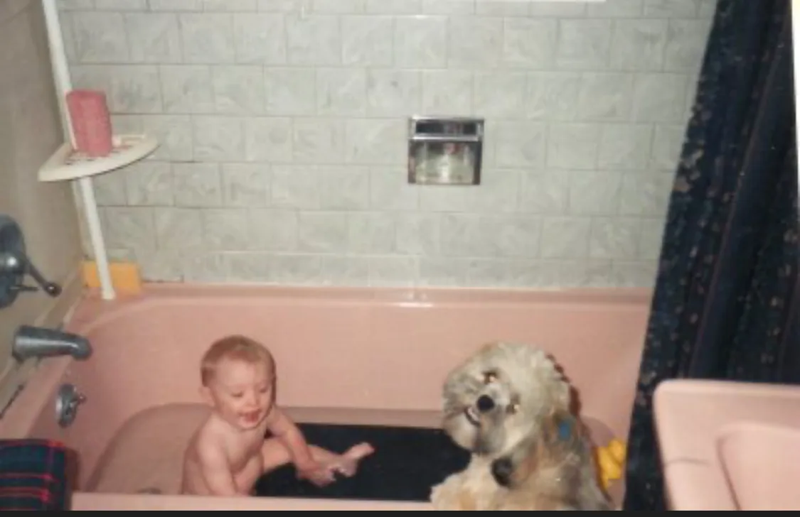A young child and a shaggy dog are sitting together in a pink bathtub filled with water. The child is smiling and splashing…